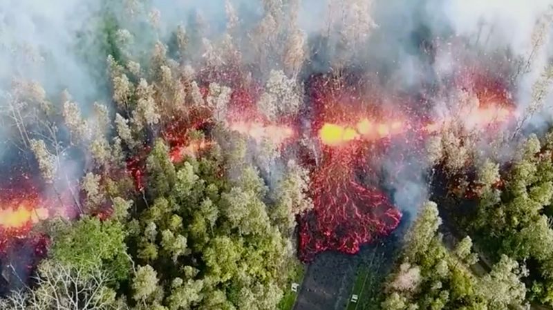 Кадр с беспилотника, показывающий разлом, образовавшийся после извержения. На фотографии видно, как из разлома на поверхность вытекает лава. Рядом с ним стоят дымящиеся деревья, а неподалеку располагаются жилые постройки. 