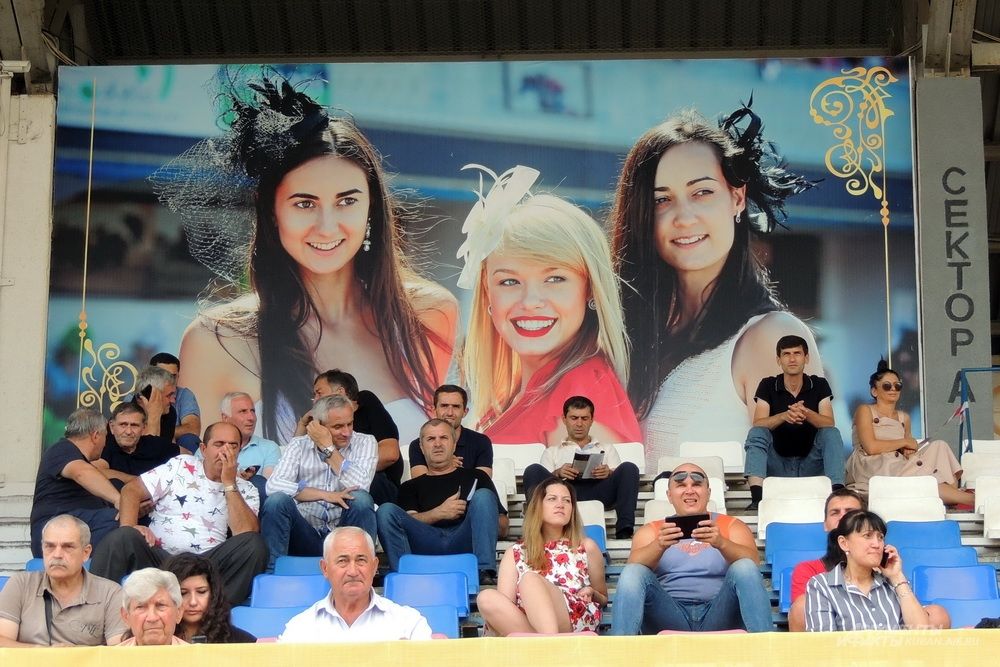 Зрители скачек на фоне баннера, напоминающего о проводимом ежегодно на Краснодарском ипподроме фестивале шляп.