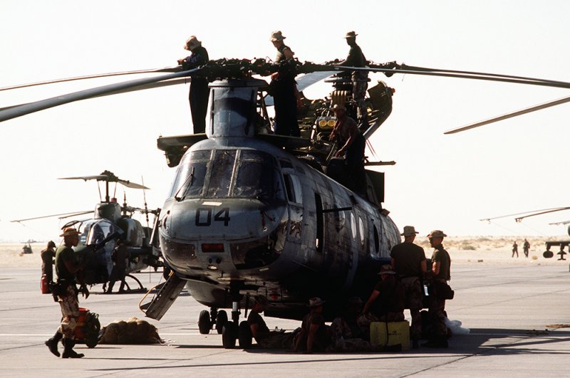 Морские пехотинцы отдыхают рядом с вертолетом CH-46 Sea Knight, пока их товарищи проверяют винты перед взлетом во время операции «Буря в пустыне».