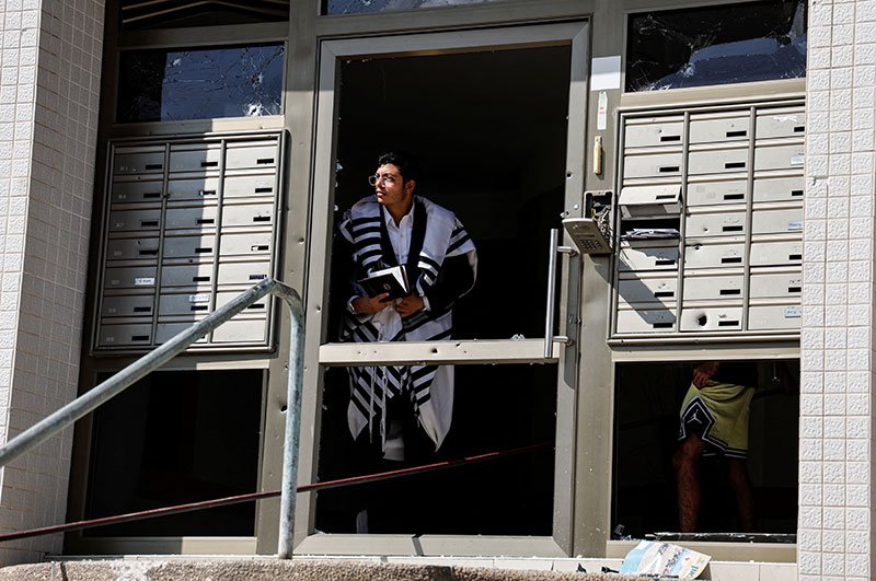 Мужчина, одетый в еврейский молитвенный платок, выглядывает из поврежденного входа в здание, когда из сектора Газа в Ашкелоне запускают ракеты.