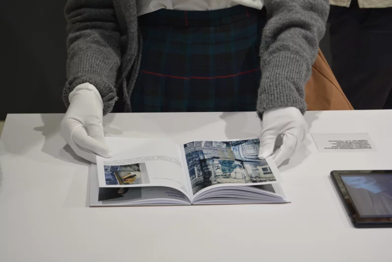 Посетительница в белых перчатках листает книгу, где собраны визуальные и текстовые материалы одного из представленных проектов. 